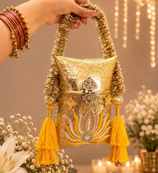 A hand wearing gold bangles holds a small ornate gold clutch bag with decorative embroidered details, a top handle, and two yellow tassels, set against a warm indoor background with flowers and hanging lights.