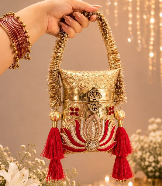 A hand wearing red bangles holds a decorative gold and red embroidered handbag with red tassels against a warm indoor backdrop with hanging lights and flowers.