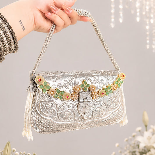 A hand holding a silver embellished evening clutch bag with floral embroidery and a beaded shoulder strap, set against a soft gray background with decorative flowers and hanging lights.