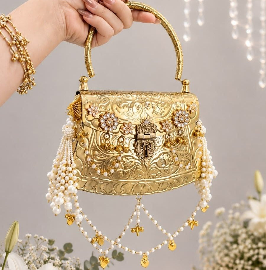 A hand holding a small ornate gold handbag decorated with intricate floral embroidery, rhinestones, and dangling pearl strands, set against a softly lit indoor background with flowers and hanging lights.