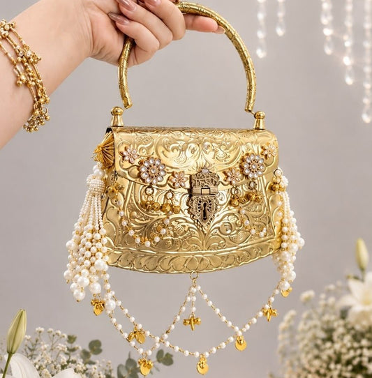 A hand holding a small ornate gold handbag decorated with intricate floral embroidery, rhinestones, and dangling pearl strands, set against a softly lit indoor background with flowers and hanging lights.