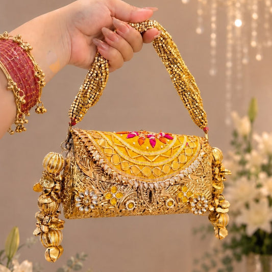 A hand wearing red bangles holds a heavily embellished gold and silver embroidered clutch purse with a decorative handle, set against a softly blurred indoor background with hanging lights and flowers.