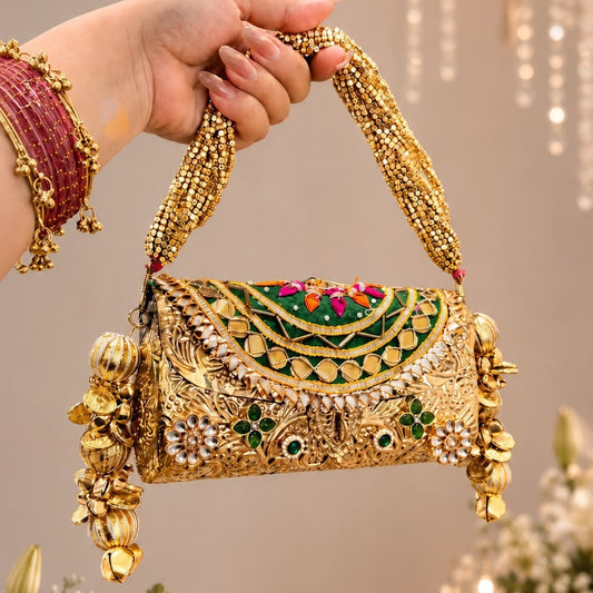 An ornate gold-toned embellished handbag with a decorative flap, colorful embroidered details, and dangling gold charms, held by a hand wearing red bangles against a softly lit indoor background with flowers and hanging lights.