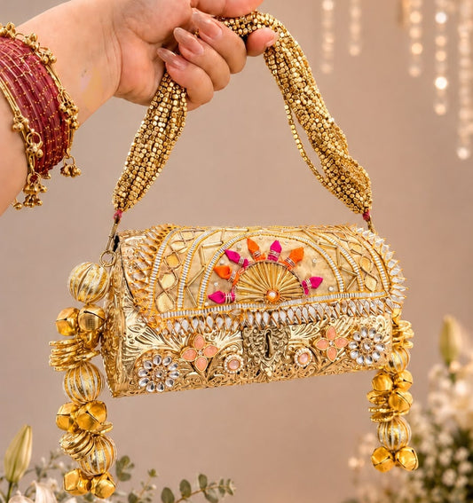 An ornate gold embellished handbag with colorful floral details and gold-toned jewelry, held by a hand wearing red bangles against a warm indoor backdrop with hanging lights and flowers.